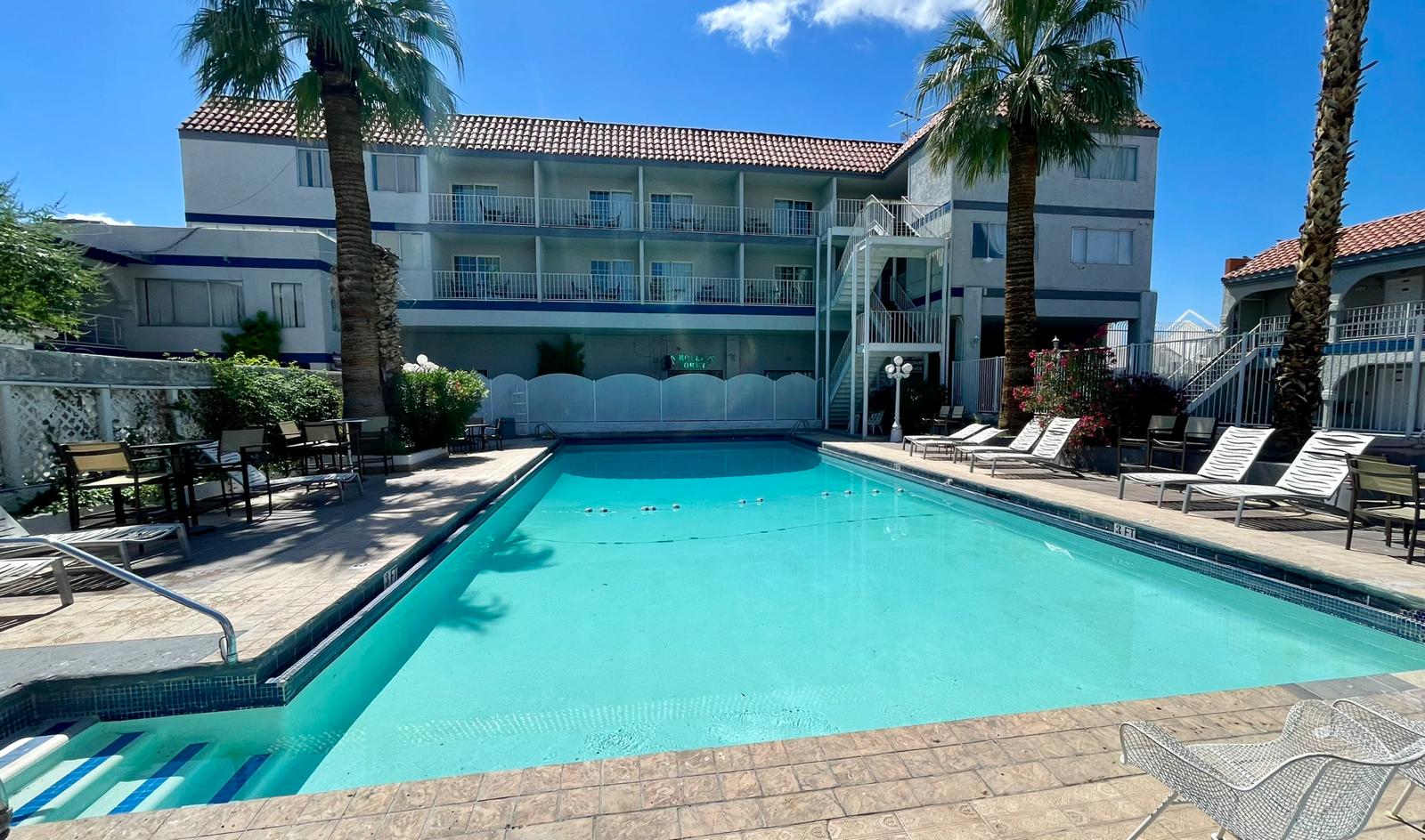 A rectangular pool with lounge chairs and palm trees in front of a multi-story hotel building.